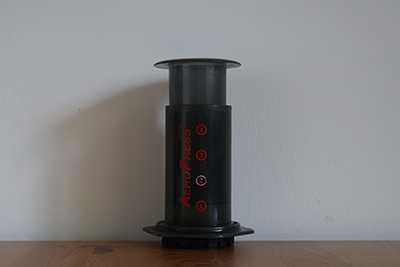 A plastic cylindrical tube with a press on the top and a red 'Aeropress' logo on the side, used to make coffee.